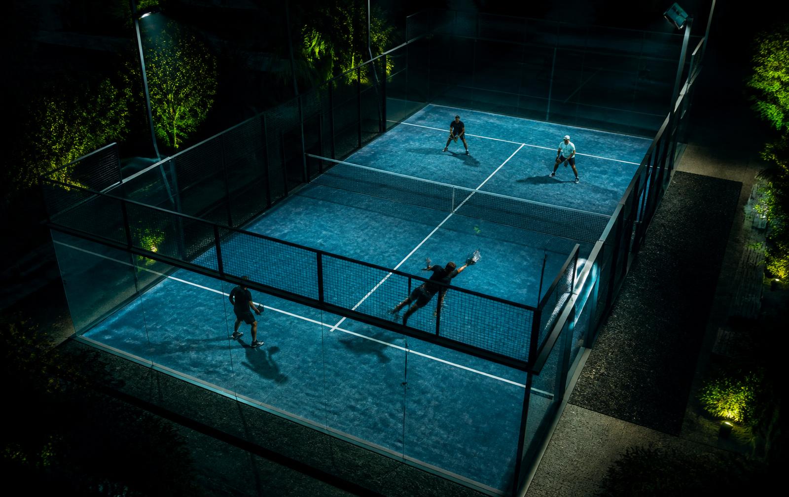Floodlit padel court at night seen from above with two pairs mid-rally